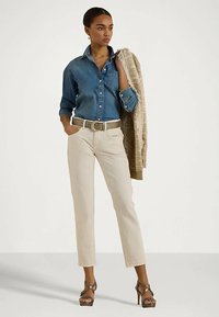 Sieviete, kura valkā zilu džinsaino kreklu, bēšas bikses ar jostu, tur bēšu adītu džemperi pār plecu un stāv uz papēžiem.