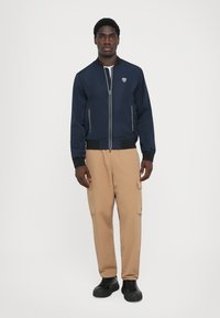 Casaco bomber azul-marinho com fecho à frente e punhos canelados, combinado com calças cargo bejes. Apresenta bolsos laterais e calçado preto.