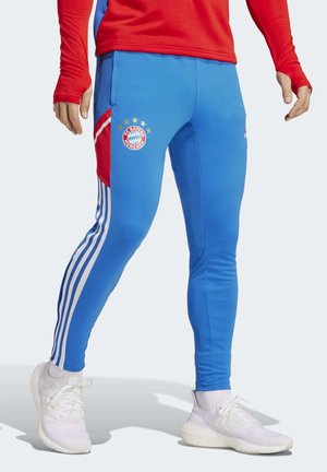 Osoba ubraną w niebieskie spodnie treningowe FC Bayern Monachium z białymi paskami Adidas i białe trampki, stojąca na jasnym tle.