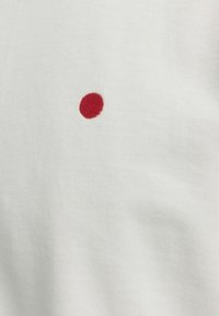 Bílý látkový tričko s malou červenou vyšívanou tečkou na přední straně. Textura je hladká a mírně vroubkovaná.