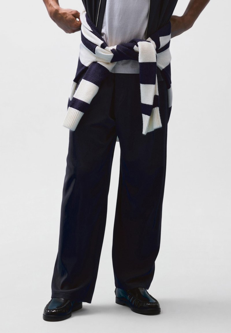 Personne portant un pantalon noir à jambes larges et des mocassins noirs, avec un pull rayé blanc et bleu marine noué autour de la taille.