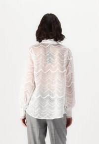 White, sheer blouse featuring a zigzag pattern with eyelet detailing and long sleeves. The fabric has a lightweight, textured finish.