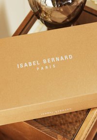 Brun papkasse med teksten "ISABEL BERNARD PARIS" i hvid. Kassen har en glat overflade og er placeret på en træoverflade.