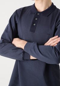 Granatowa koszulka polo z długim rękawem, kołnierzykiem, trzema guzikami oraz subtelnym haftem inicjałów "MN" na piersi. Gładka faktura tkaniny.