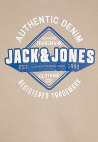Bež pamukasta majica s plavim i bijelim grafičkim dizajnom s natpisom "JACK & JONES" i "AUTHENTIC DENIM" unutar dijamantskog oblika.