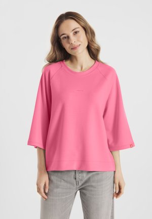 Vrouw met golvend haar, gekleed in een losse roze longsleeve en lichtgrijze spijkerbroek, staand tegen een effen lichte achtergrond.