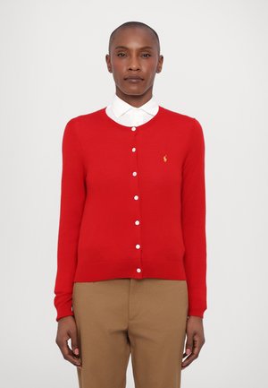 Punainen neulecardigan näyttävillä valkoisilla napeilla, pyöreä kaula-aukko ja pieni brodattu logo. Pukeutunut valkoisen kauluspaidan päälle, yhdessä beigenhousujen kanssa.