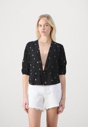 Svart kort blouse med diamantmønster, dyp utringning, korte puffarmar og knytning i midjen. Kombinert med hvite slitte shorts.
