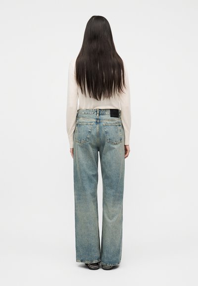 Γυναίκα με μακριά σκούρα μαλλιά που φορεί ανοιχτό μπεζ μακρυμάνικη μπλούζα και φαρδιά ξεθωριασμένα μπλε τζιν, στέκεται γυρισμένη πλάτη πάνω σε λευκό φόντο.