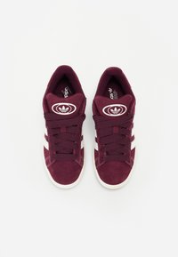 Kastanienbraune Wildledersneaker mit weißen Streifen, rundem Logo-Patch auf der Zunge, traditionellem Schnürdesign und strukturierten Stoffakzenten.