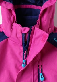 Détail d'une veste imperméable rose avec une fermeture éclair dotée de tirettes turquoise, col ajustable et accents bleu marine foncé sur les épaules et les poignets.