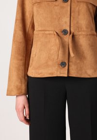 Bruine faux-suède jas met een strik taille, knoopsluiting en twee voorzakken. Zwarte, op maat gemaakte broek eronder. Glad textuur.