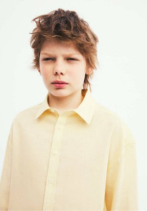 Garçon aux cheveux bruns ébouriffés portant une chemise jaune clair à boutons, regardant devant lui avec une expression neutre sur un fond uni.