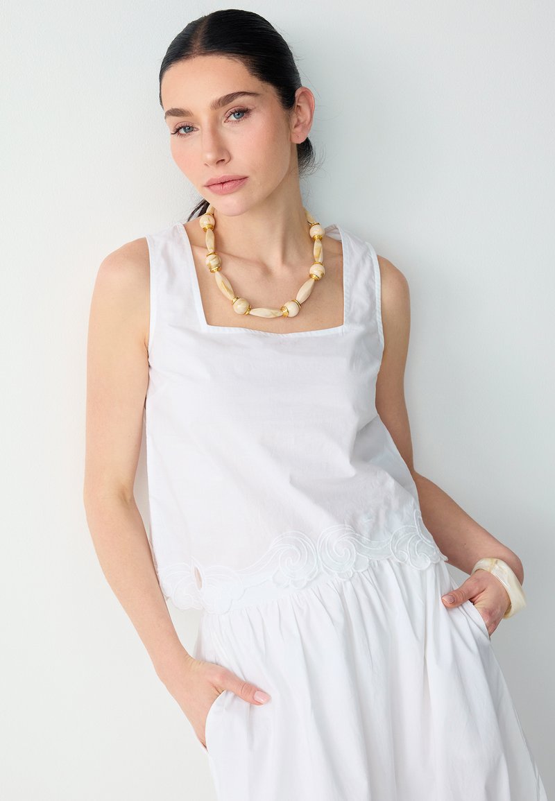 Femme portant un haut blanc sans manches à ourlet festonné, une jupe blanche, un collier de perles beige et un bracelet beige, posant les mains dans les poches.