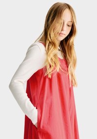 Abito rosso senza maniche con due tasche laterali, indossato sopra una maglietta bianca a maniche lunghe. Tessuto morbido con una vestibilità comoda e una leggera lucentezza.