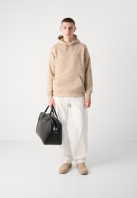 Beige hoodie with kangaroo pocket, white trousers, tan boots, and a structured black leather bag with dual handles. Minimalistic design.