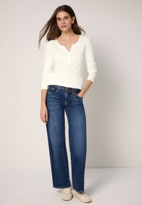 Witte henley top met lange mouwen en een V-hals met inkeping; gecombineerd met wijde blauwe denim jeans; model draagt witte sneakers.