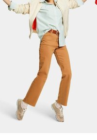 Personne portant un pantalon beige, des baskets beiges, des chaussettes à rayures, une chemise à rayures claire et une veste légère, debout jambes écartées sur un fond blanc.