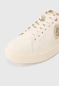 Bijela tenisica od kože s glatkim gornjim dijelom, okruglim vrhom, ravnim vezicama i zlatnim detaljem. Ima teksturiranu gumenastu potplat i blistave ukrase.