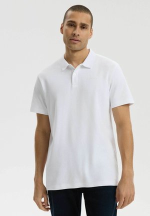 Młody mężczyzna stoi, ubrany w gładką białą koszulkę polo z krótkim rękawem i ciemne spodnie na neutralnym tle.