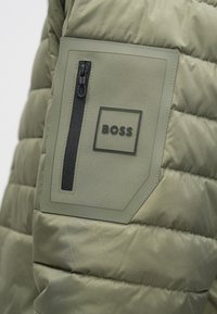 Detail rukáva olivovo zelenej prešívanej bundy s čiernym vreckom na zips a štvorcovou nášivkou s nápisom "BOSS".