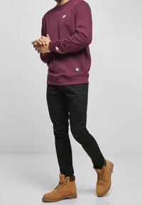 Hombre vestido con un sudadera color burdeos, pantalones ajustados negros y botas de trabajo beige con cordones, de pie frente a un fondo claro y simple.