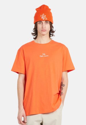 Camiseta de algodón naranja de manga corta con cuello redondo y logo bordado en blanco "Polo Ralph Lauren". Combinada con un gorro naranja a juego.