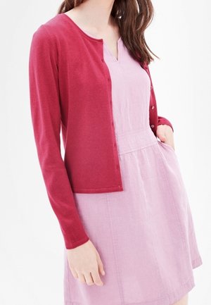Femme portant une robe sans manches rose clair avec un cardigan boutonné rose foncé et une main dans la poche de la robe.