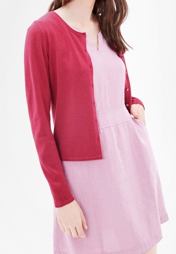 Femme portant une robe sans manches rose clair avec un cardigan boutonné rose foncé et une main dans la poche de la robe.