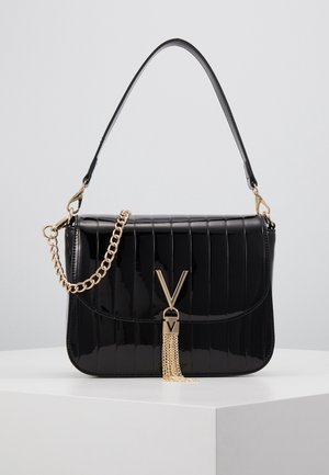 Sac à main noir brillant avec un design cousu verticalement, accent chaîne dorée et détail pompon doré en forme de "V" sur le rabat avant, posé sur une surface blanche.
