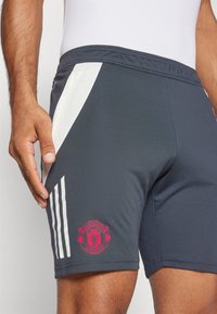 Grå träningsshorts med vita sidopaneler, utrustade med en rosa Manchester United-logotyp på nedre vänstra benet och tre vita ränder.