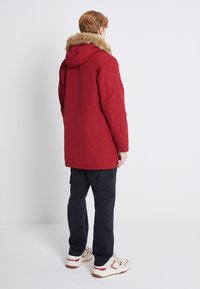 Röd parkajacka med fuskpäls på huvan, strömlinjeformad design och lång nederkant. Kombinerad med svarta cargobyxor och vita sneakers med röda detaljer.