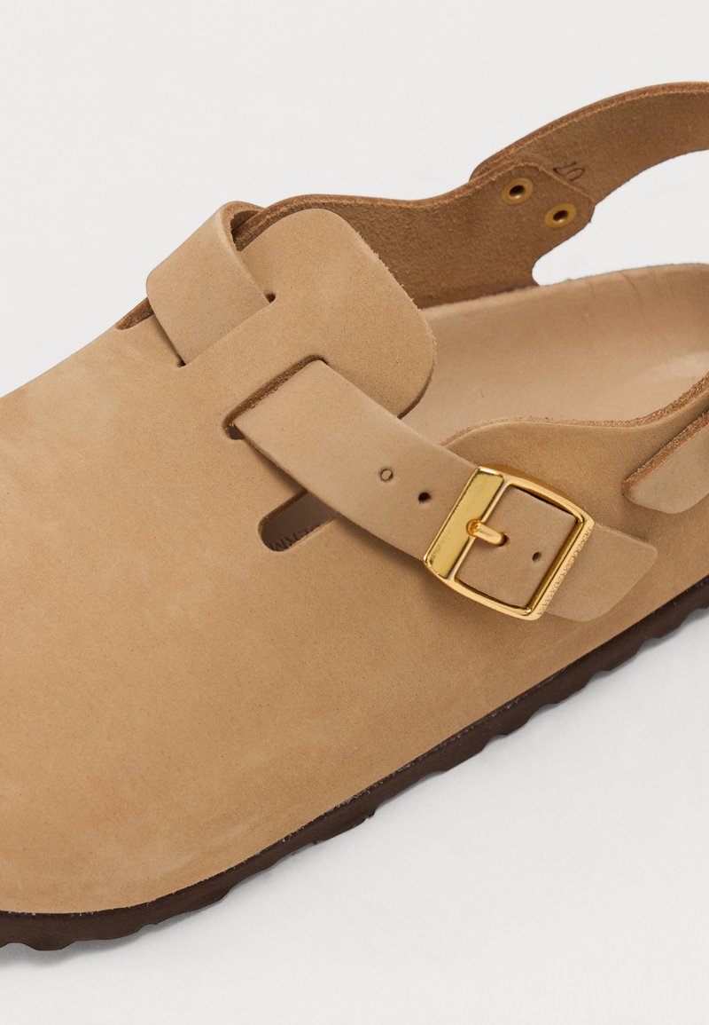 Sandalo in pelle color tan con superficie texturizzata, dotato di una fibbia in metallo dorato e design aperto sul retro. La suola è marrone e leggermente scanalata.