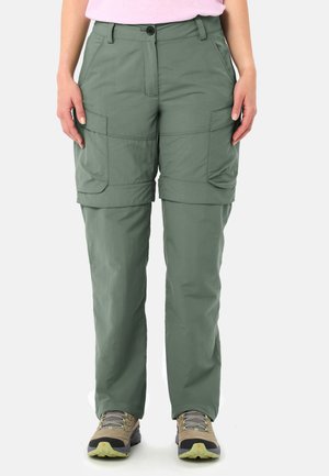 Femme portant un pantalon cargo convertible vert olive et des chaussures de randonnée, debout, mains détendues le long du corps.