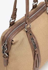 Beżowa torba płócienna z brązowymi skórzanymi akcentami, z podwójnymi uchwytami i srebrnym zamkiem błyskawicznym. Charakteryzuje się teksturowaną powierzchnią i szczegółowym przeszyciem.