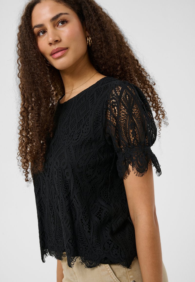 Femme aux cheveux bouclés portant un haut en dentelle noir à manches courtes et un pantalon beige, avec des boucles d'oreilles créoles en or et un collier délicat.