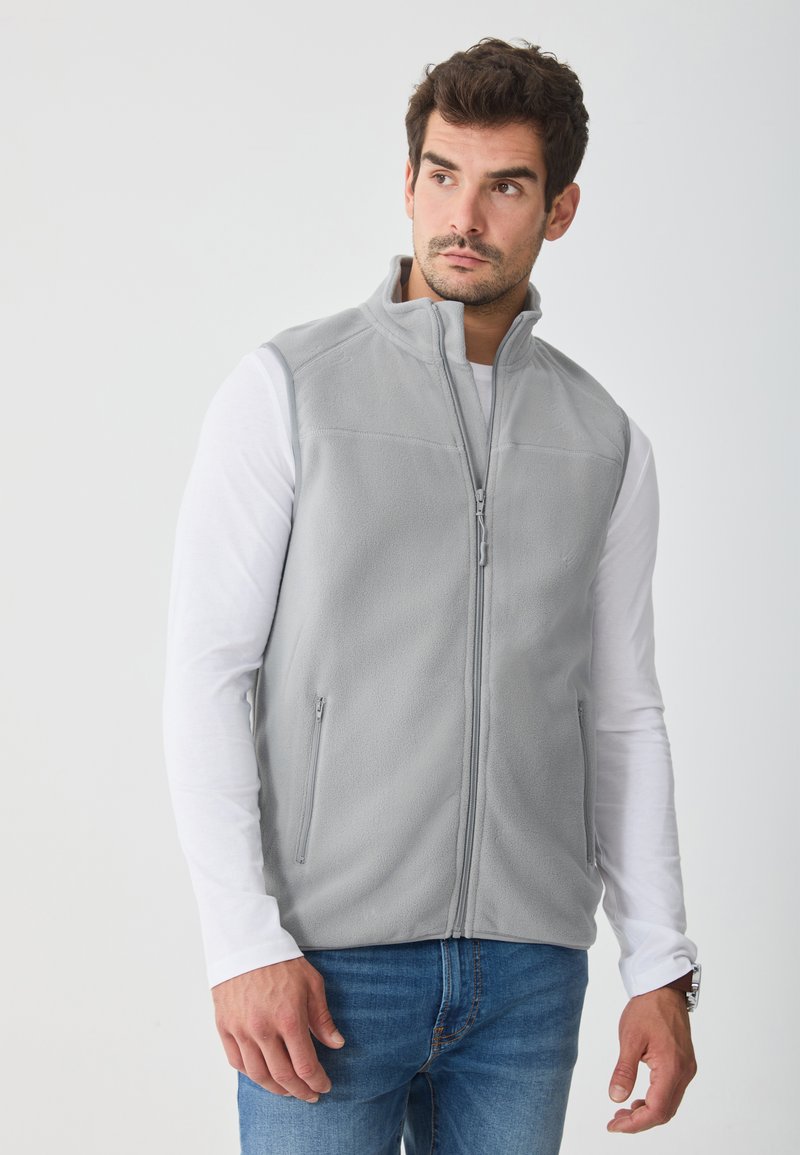Vest gri din fleece cu guler înalt, fermoar frontal și două buzunare laterale, purtat peste o cămașă albă cu mânecă lungă și blugi albaștri.