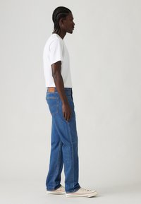White short-sleeve t-shirt paired with blue denim jeans, featuring a straight cut, classic five-pocket design, and brown leather patch on the back.