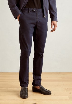 Mężczyzna ubrany w ciemnoniebieskie, dopasowane spodnie, czarne mokasyny i dopasowaną marynarkę w tym samym odcieniu granatu, stojący na jasnej drewnianej podłodze.