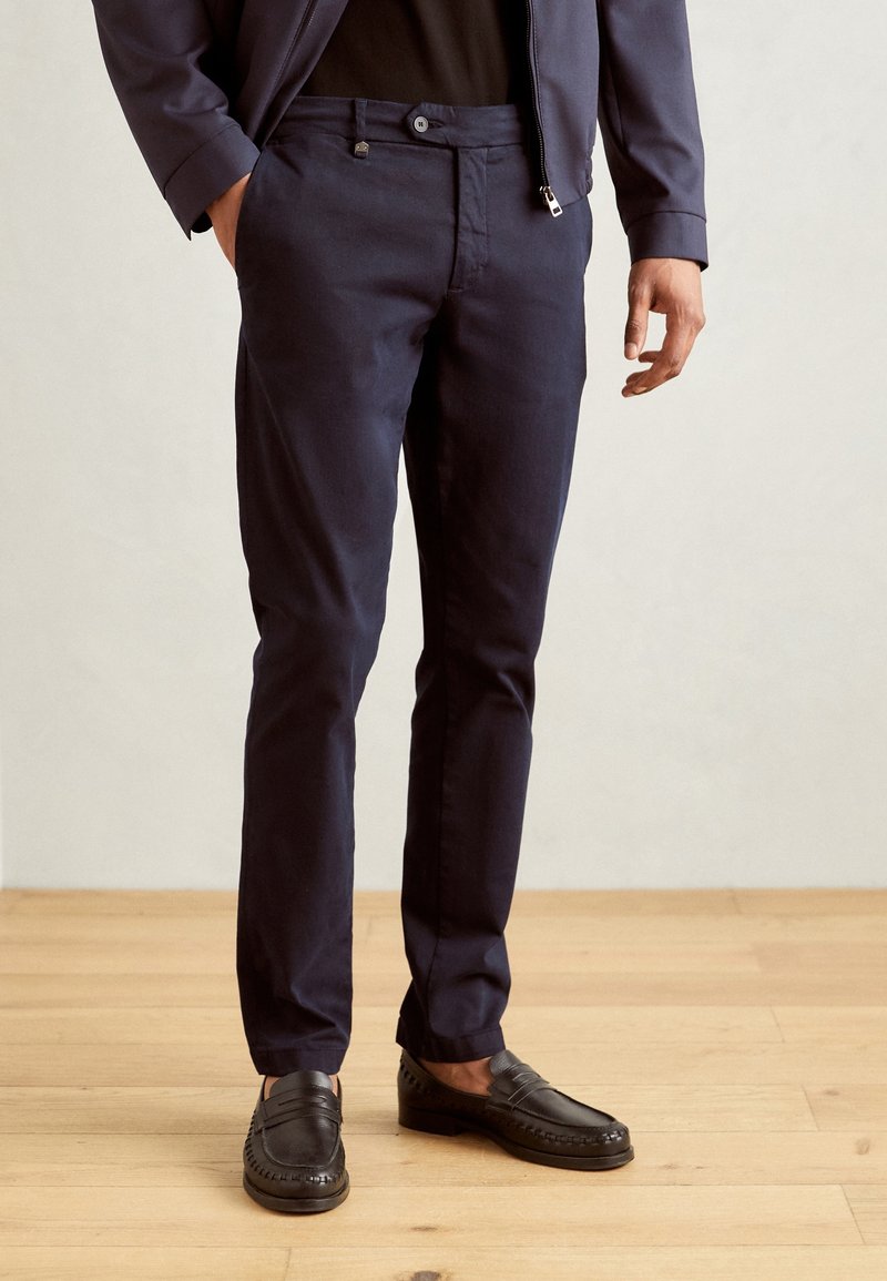 Homme portant un pantalon slim bleu marine foncé, des mocassins noirs et une veste assortie bleu marine, debout sur un sol en bois clair.