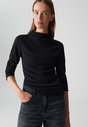 Junge Frau mit glattem Haar trägt ein schwarzes, geripptes Langarmoberteil und dunkel gewaschene Jeans, mit einer Hand in der Tasche vor einem hellen Hintergrund.
