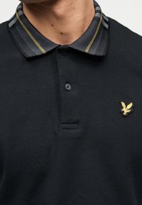 Άντρας που φοράει μαύρο πόλο πουκάμισο με υφασμένο γιακά που έχει γκρι και κίτρινες ρίγες και ένα μικρό κίτρινο λογότυπο αετού στο στήθος.