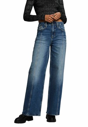 Niebieskie dżinsy o szerokich nogawkach z dwoma pionowymi szwami. Tkanina w średnim odcieniu z gładką fakturą, zestawiona z czarnymi masywnymi butami.