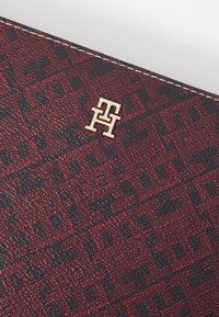 Materiał w czerwono-czarny wzór o teksturowanej powierzchni, z akcentem złotego logo. Białe przeszycia podkreślają krawędź.