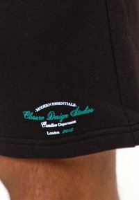 Μαύρο ύφασμα με ραμμένο κείμενο "Modern Essentials. Closure Design Studios Creative Department London 2015" κοντά στο στρίφωμα.