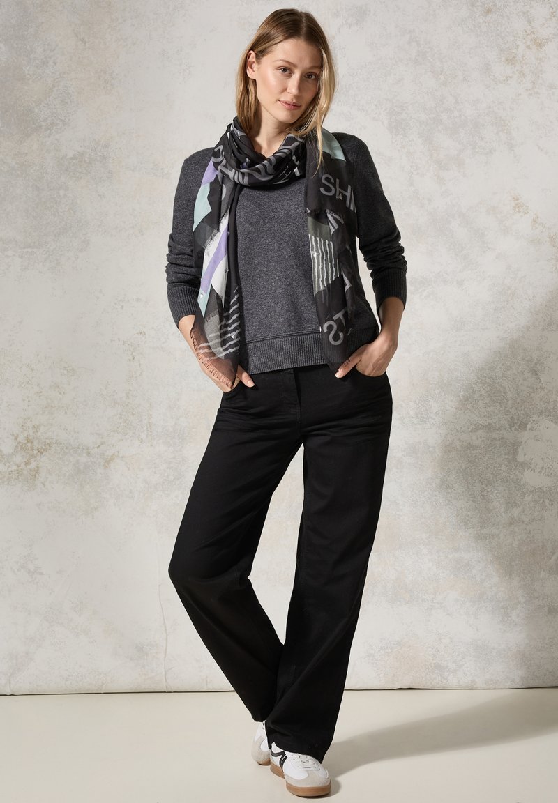 Maglione grigio con finitura testurizzata, pantaloni larghi neri e una sciarpa fantasia con forme geometriche e colori tenui. Sneakers casual.