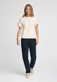 Camiseta de manga corta crema con un logo cuadrado en el pecho, combinada con pantalones marinos y zapatos de color claro, de pie frente a un fondo liso.