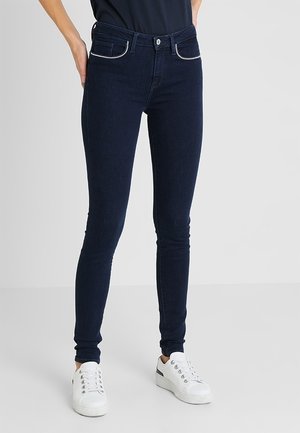 Person iført mørkeblå skinny jeans med hvide syninger og hvide sneakers, stående mod en ensfarvet lys grå baggrund.