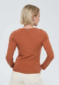 Femme aux cheveux blonds courts portant une chemise à manches longues couleur rouille et un pantalon écru, de dos avec les mains sur les hanches.