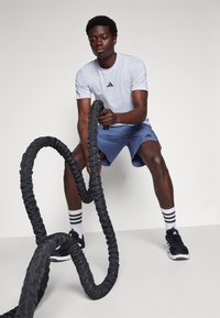 Black battle rope held by a person in a light gray Adidas shirt, blue shorts, and black athletic shoes with white striped socks.